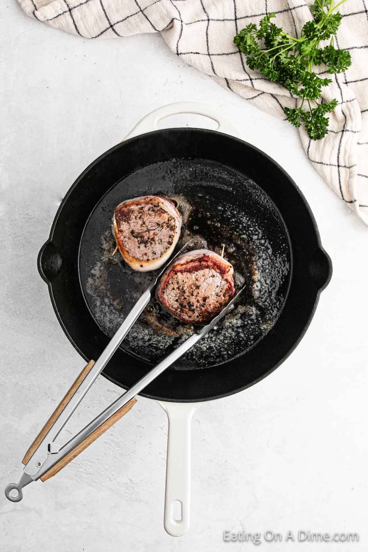 A frying pan sizzles with bacon-wrapped filet mignon steaks, as tongs hold one juicy piece above the pan. Fresh parsley adds a touch of green next to a checked kitchen towel on the light-colored countertop, completing the culinary scene.