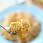 A close-up of a fork holding a bite of creamy, cheesy risotto made with the easy cheesy cauliflower rice recipe, garnished with chopped herbs, with a blurred background of the dish on a plate.