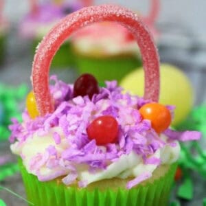 Discover this Easy Easter Cupcakes Recipe: A cupcake in a green wrapper, topped with white frosting, purple coconut shreds, colorful candy eggs, and a pink gummy handle to mimic an Easter basket. Surrounding the cupcake is bright green decorative grass for an extra festive flair.