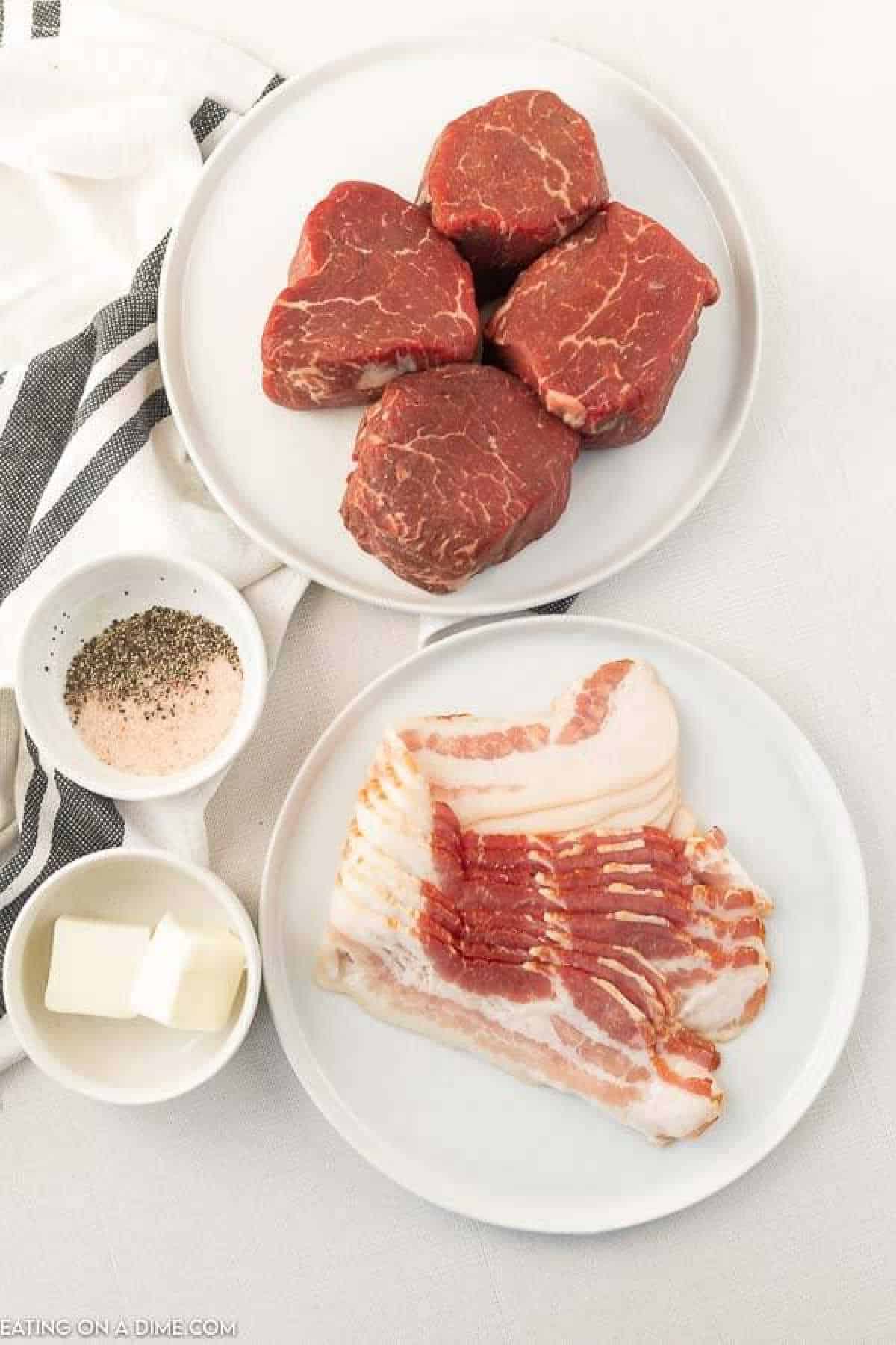 A top-down view of four raw filet mignon steaks on a white plate, raw bacon strips ready to turn them into bacon-wrapped filet on a separate white plate, a small dish of butter, and two small bowls containing coarse salt and black pepper. A striped cloth napkin is partially visible underneath the plates.