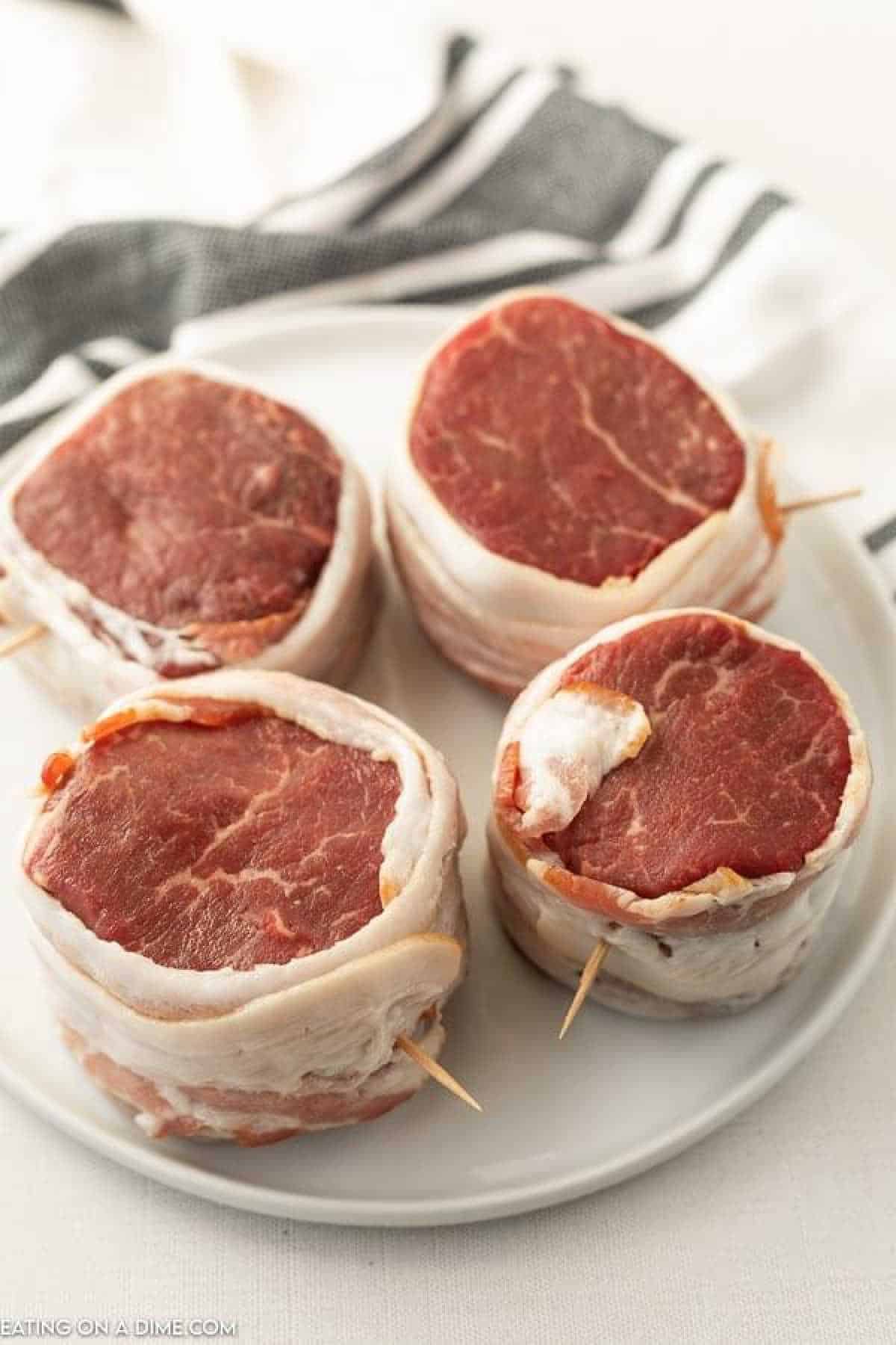 A white plate contains four Bacon Wrapped Filet mignon steaks, each secured with toothpicks. A black and white striped cloth is partially visible in the background on a white surface.