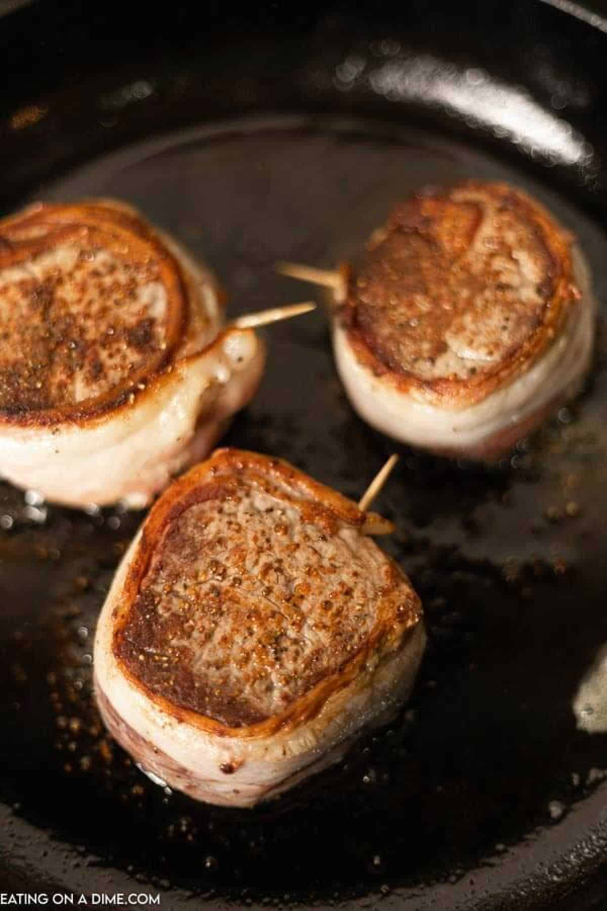 Three bacon-wrapped filet mignon steaks sizzle in a black skillet, each steak browning beautifully with visible toothpicks holding the bacon securely in place. The rustic skillet adds a charming touch to the delectable cooking process.