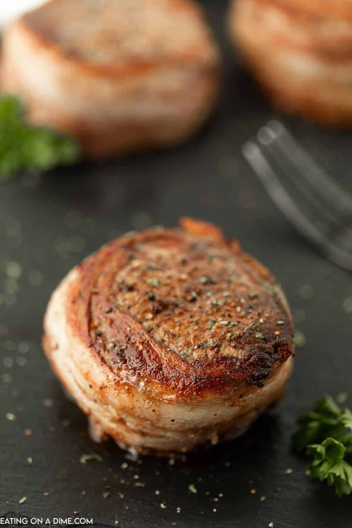 Close-up of a Bacon Wrapped Filet mignon garnished with herbs on a dark surface. The filet appears juicy and well-seasoned, with a fork and another filet mignon slightly blurred in the background. Fresh parsley is also visible on the surface.