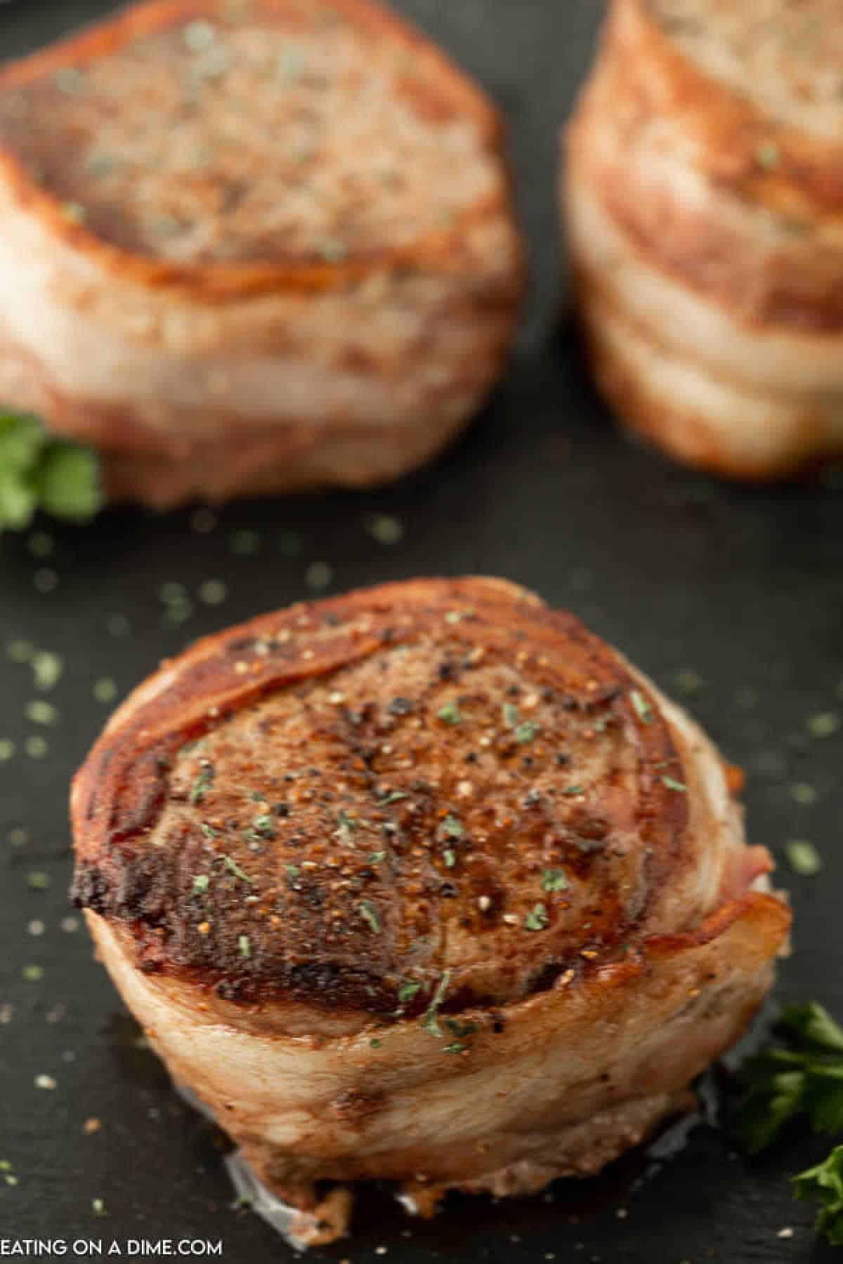 Close-up of bacon-wrapped filet mignon steaks cooking on a dark surface, garnished with herbs and a few sprigs of parsley in the background. The bacon wrapped filets are browned and crisp on the outside, indicating they are seared to perfection.