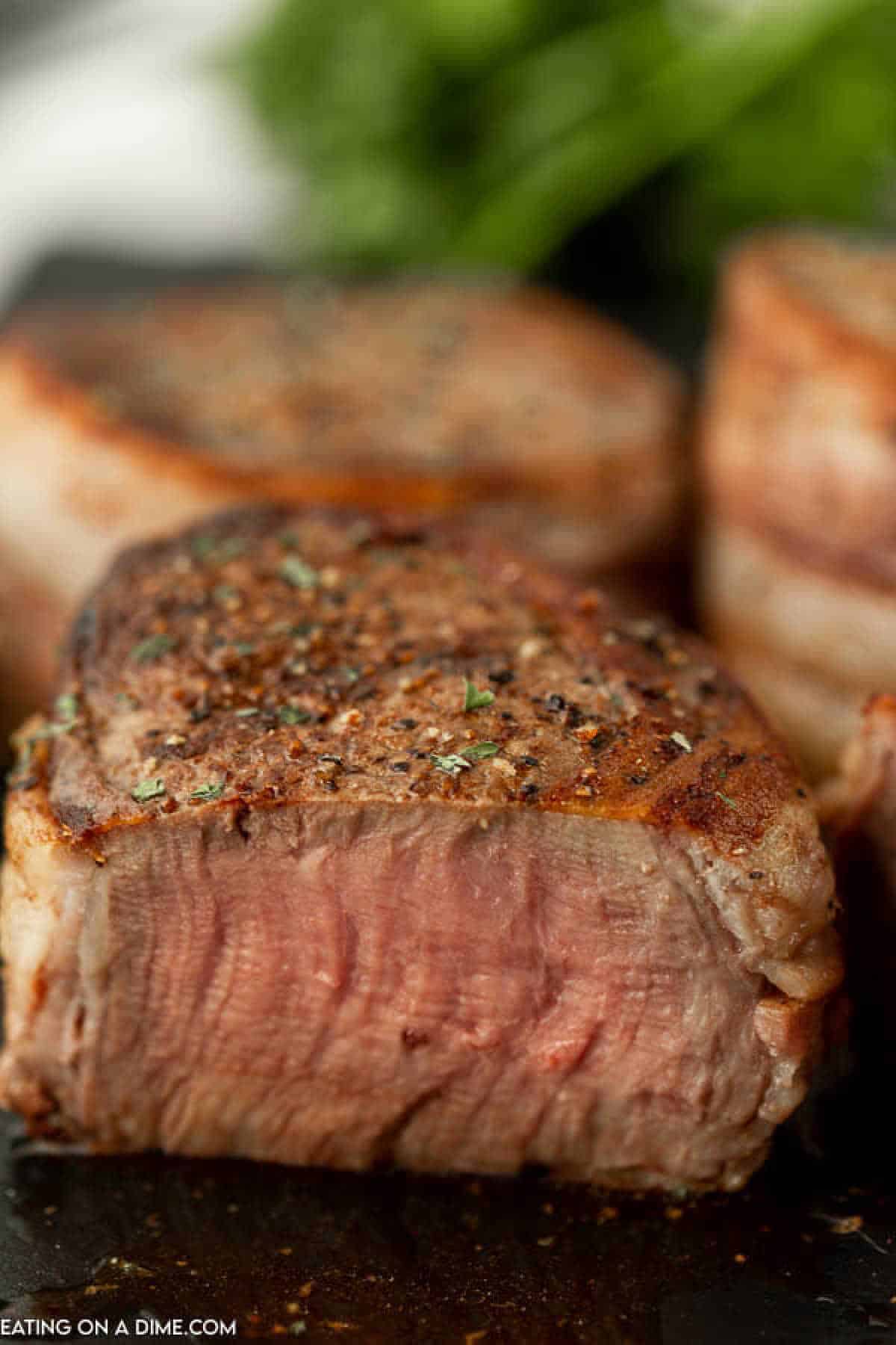 Close-up of a perfectly cooked, medium-rare bacon wrapped filet, showcasing its juicy interior and seasoned, seared exterior. The steak is garnished lightly with herbs, and there is a blurred green garnish in the background.