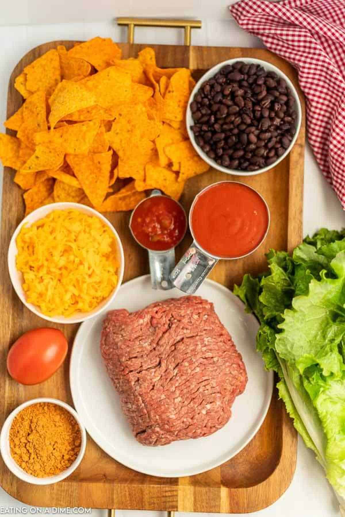 A wooden tray displays ingredients for a Doritos Taco Salad: ground beef, tortilla chips, black beans, shredded cheese, salsa, tomato sauce, a whole tomato, taco seasoning, and lettuce. A red and white checkered cloth sits beside the tray.