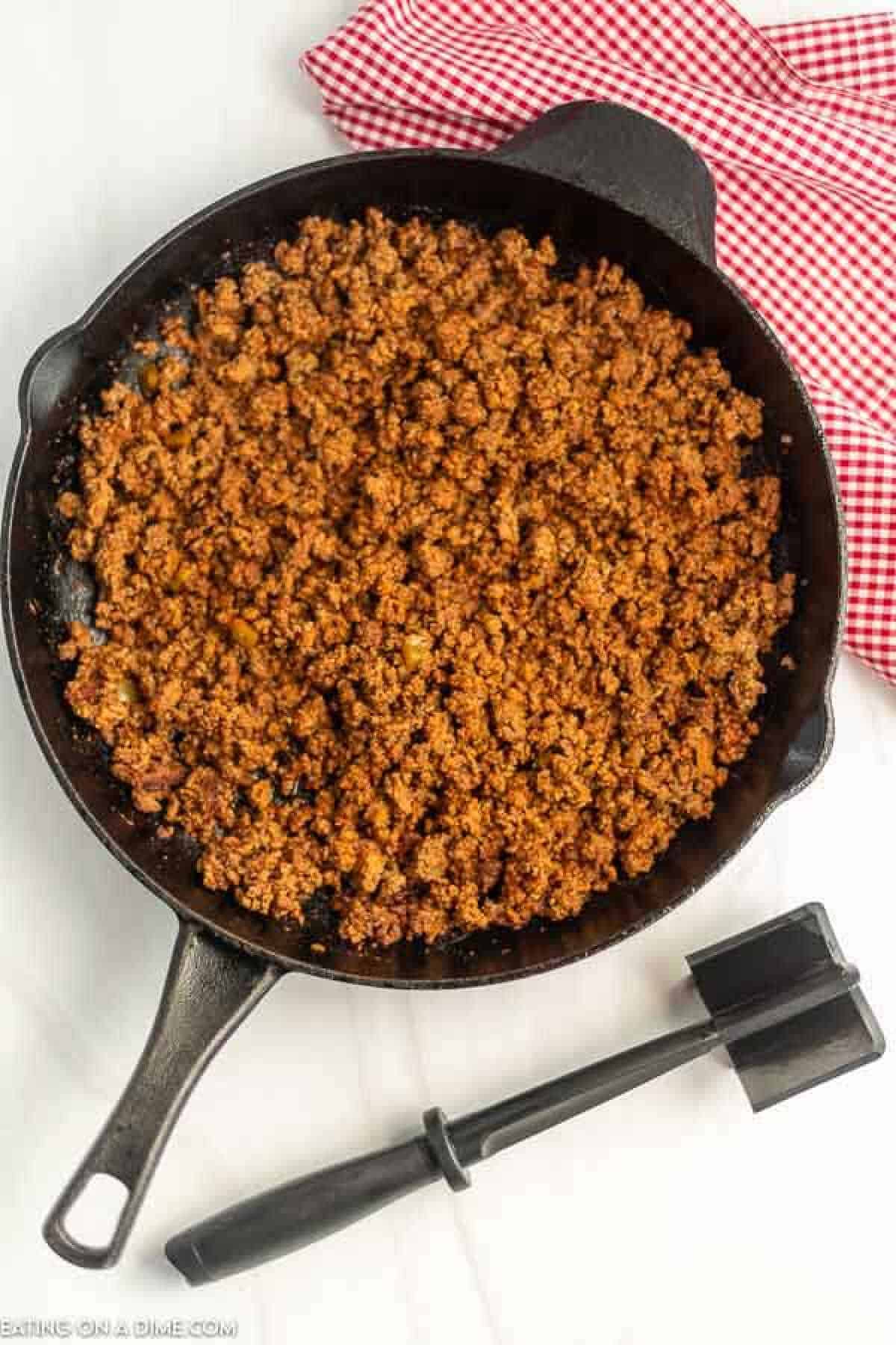 A cast iron skillet filled with browned ground beef, reminiscent of a Doritos Taco Salad, sits on a white surface. A black spatula rests nearby, and a red and white checkered cloth is partially visible in the background.