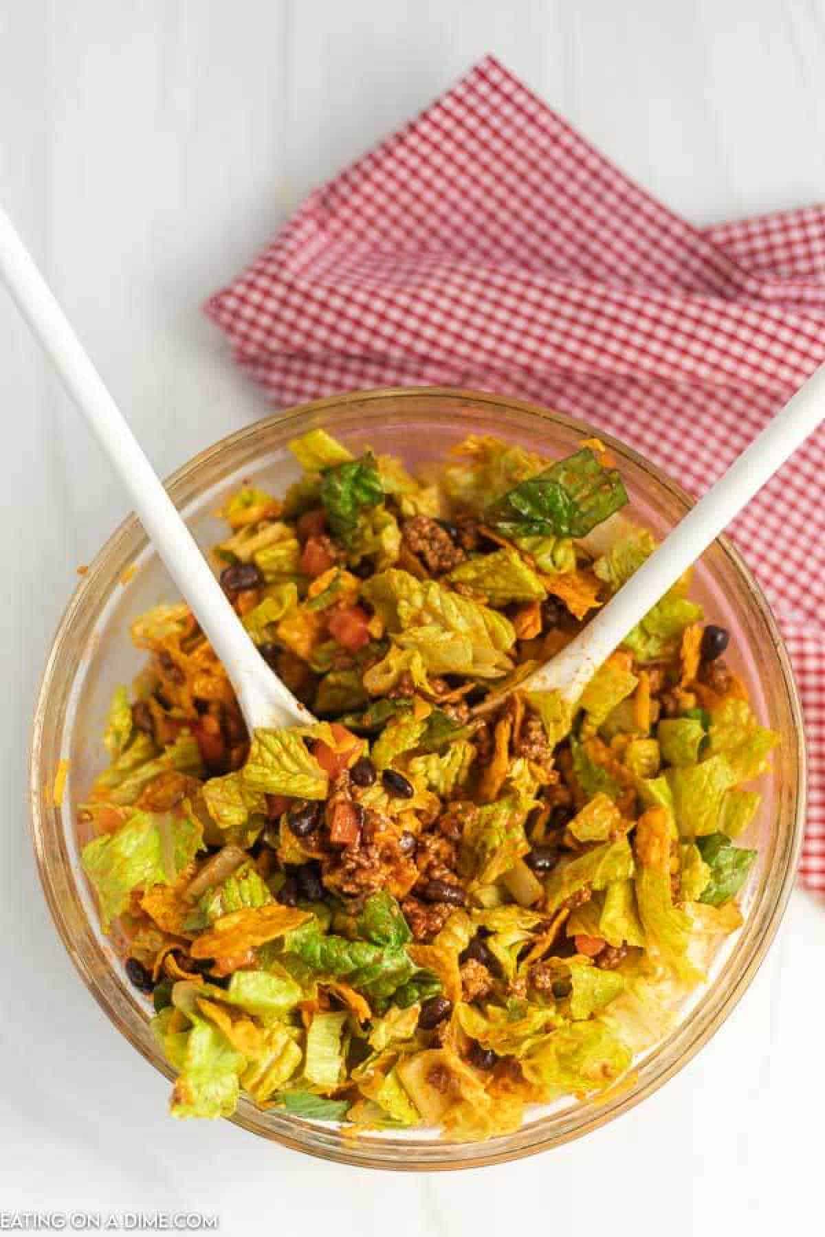 A glass bowl brims with Doritos Taco Salad featuring crisp lettuce, black beans, ground beef, and diced tomatoes. Two white salad servers rest in the mix, while a folded red and white checkered cloth is placed on the white surface in the background.