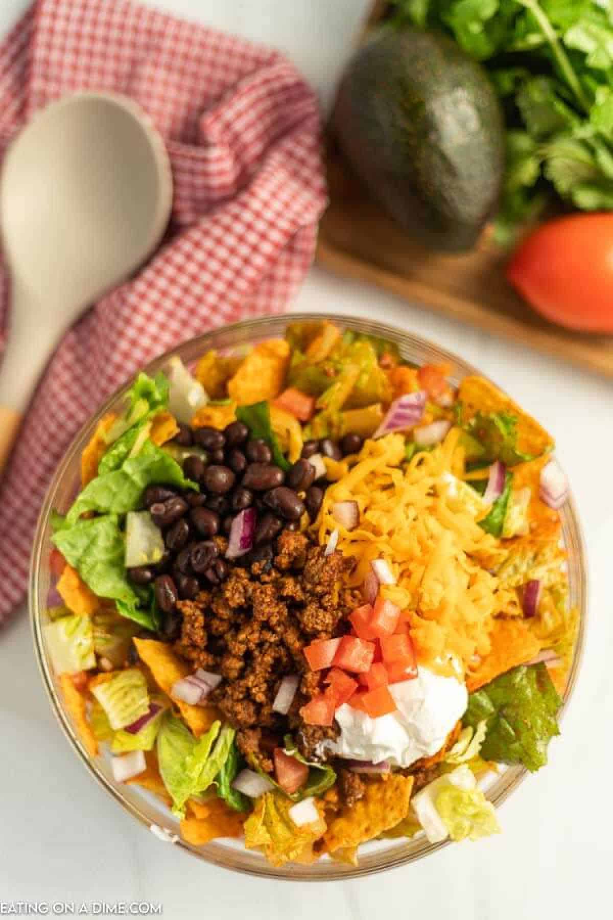 A bowl of Doritos taco salad features seasoned ground beef, crunchy Doritos, black beans, shredded cheese, chopped tomatoes, red onions, crisp lettuce, and a dollop of sour cream. Nearby sit a wooden spoon, checked cloth, avocado, tomato, and fresh cilantro.