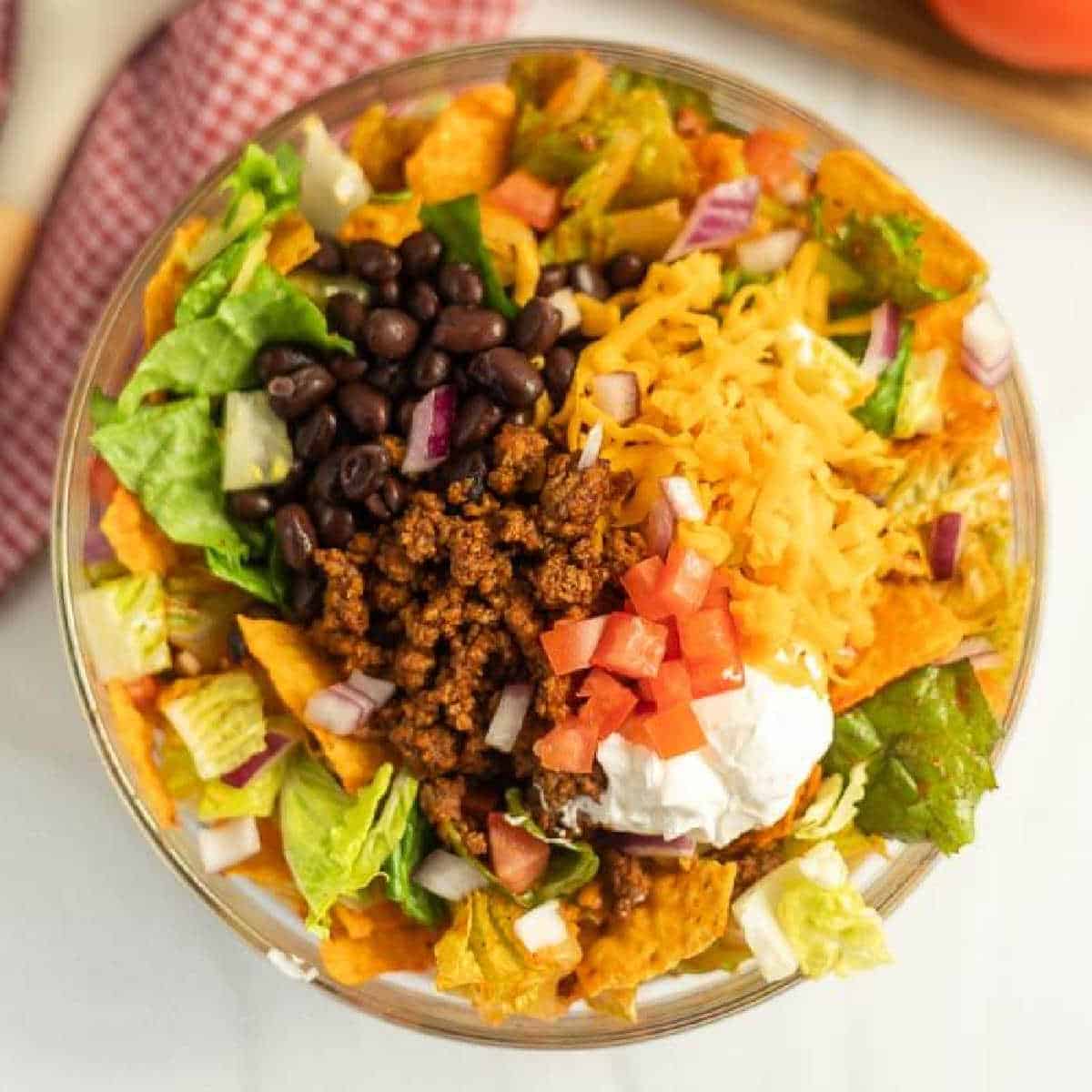 A vibrant Doritos Taco Salad in a clear bowl features seasoned ground beef, black beans, shredded cheddar cheese, chopped tomatoes, diced onions, lettuce, and a dollop of sour cream on top. Crushed Doritos are mixed in for an irresistible crunch.