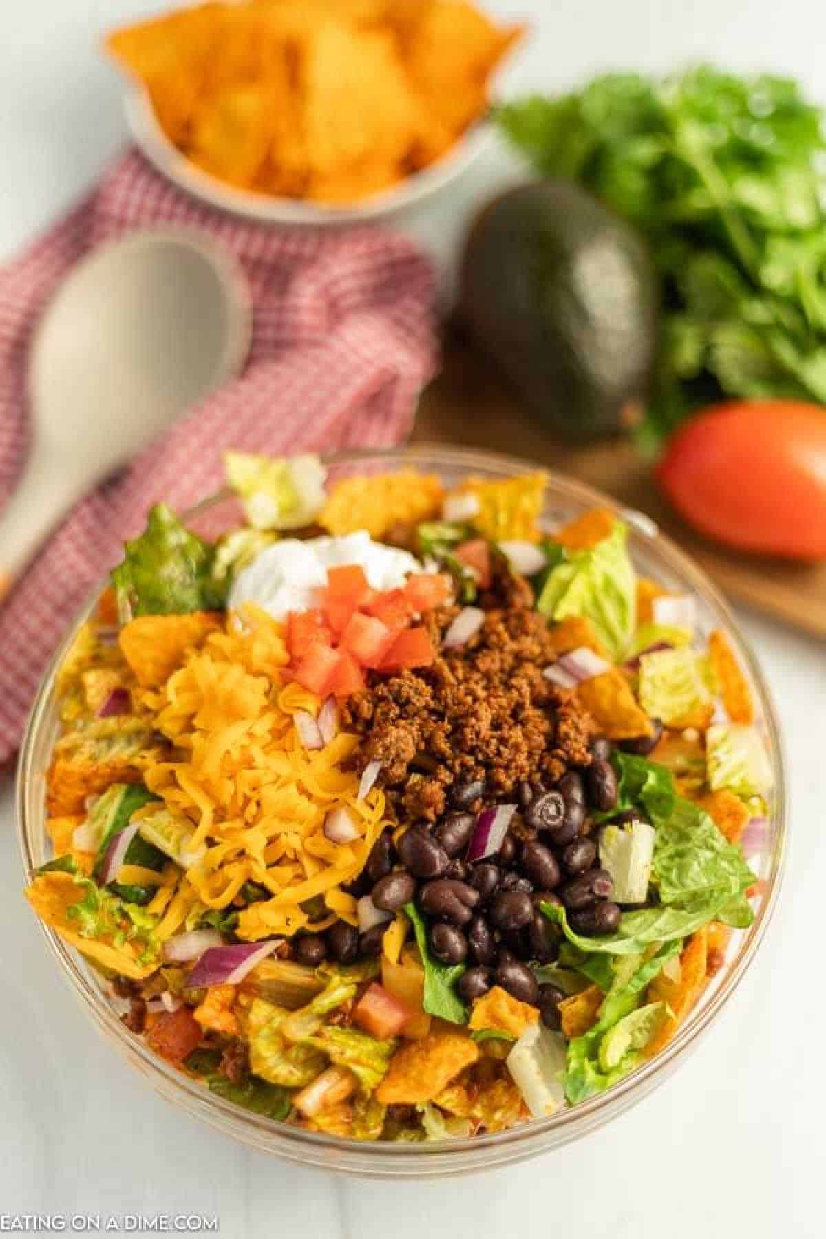 A bowl of Doritos taco salad with ground meat, black beans, shredded cheese, lettuce, diced tomatoes, sour cream, and crumbled tortilla chips. In the background, a whole avocado, tomato, and fresh cilantro await. A red checkered napkin sits charmingly beside the bowl.