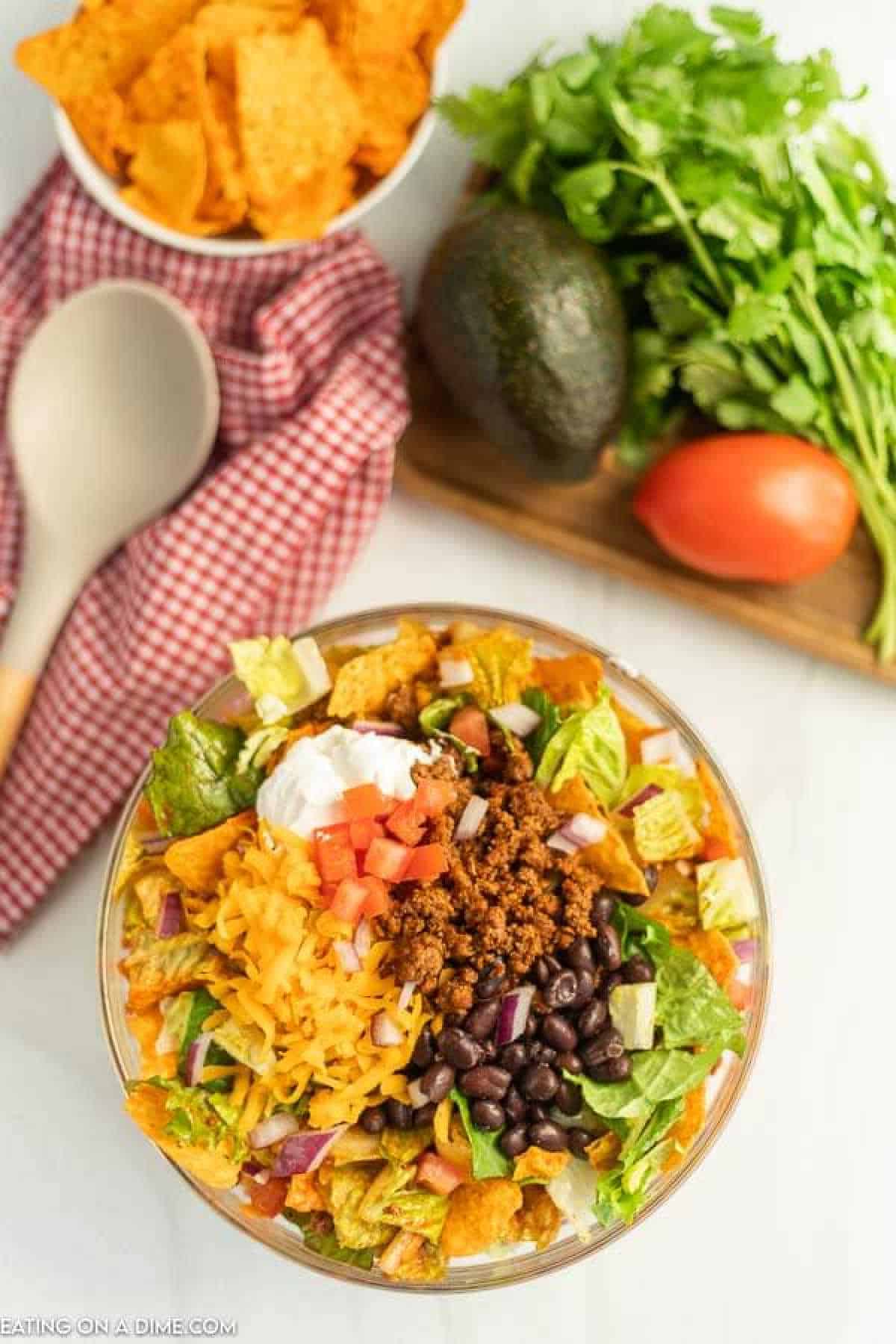 A Doritos Taco Salad in a glass bowl features layers of lettuce, seasoned ground beef, black beans, shredded cheese, diced tomatoes, and a dollop of sour cream. Nearby are tortilla chips, avocado, tomato, cilantro, and a wooden spoon for serving.