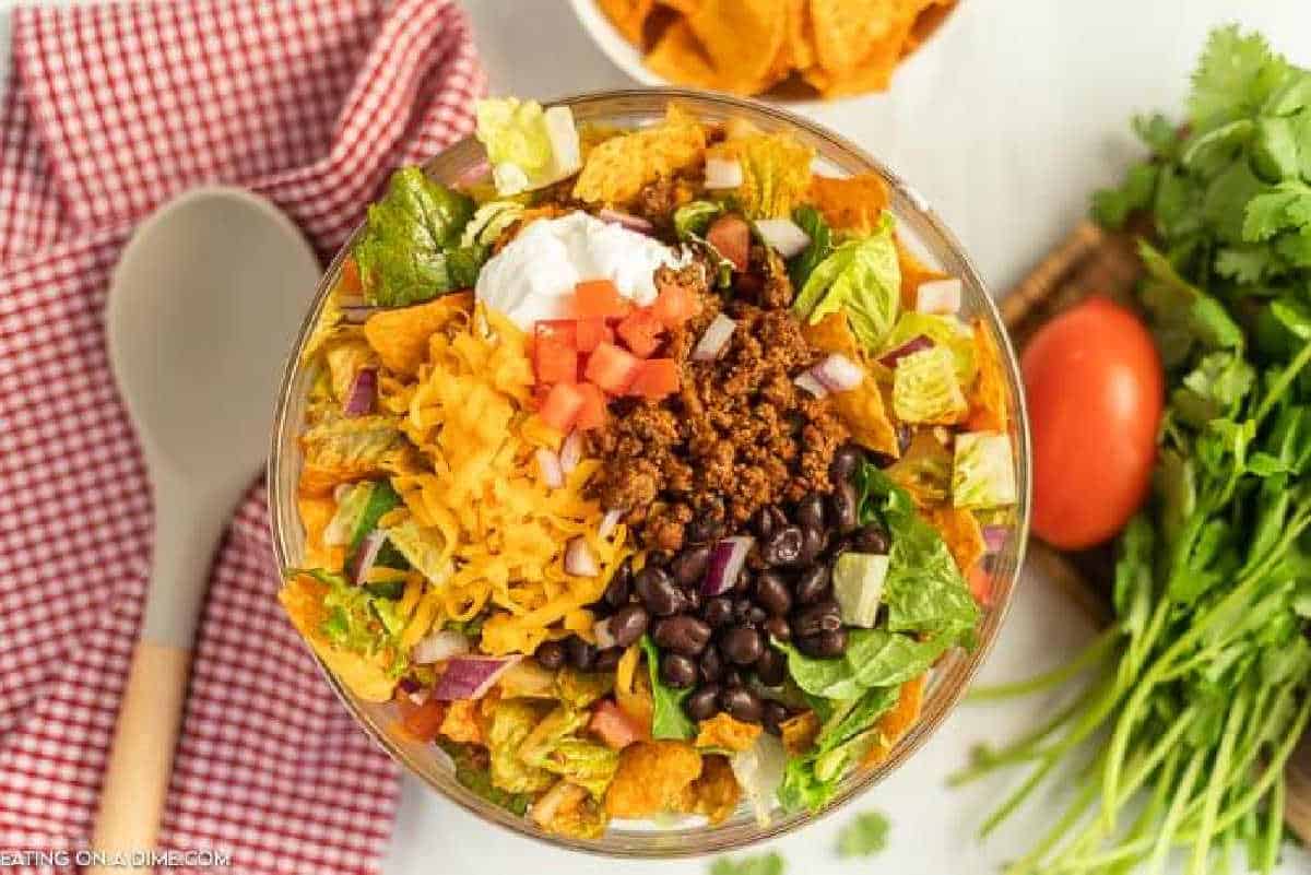 A bowl of Doritos taco salad featuring ground beef, black beans, shredded cheddar cheese, diced tomatoes, lettuce, sour cream, and tortilla chips. A red checkered cloth, serving spoon, and fresh cilantro are nearby.