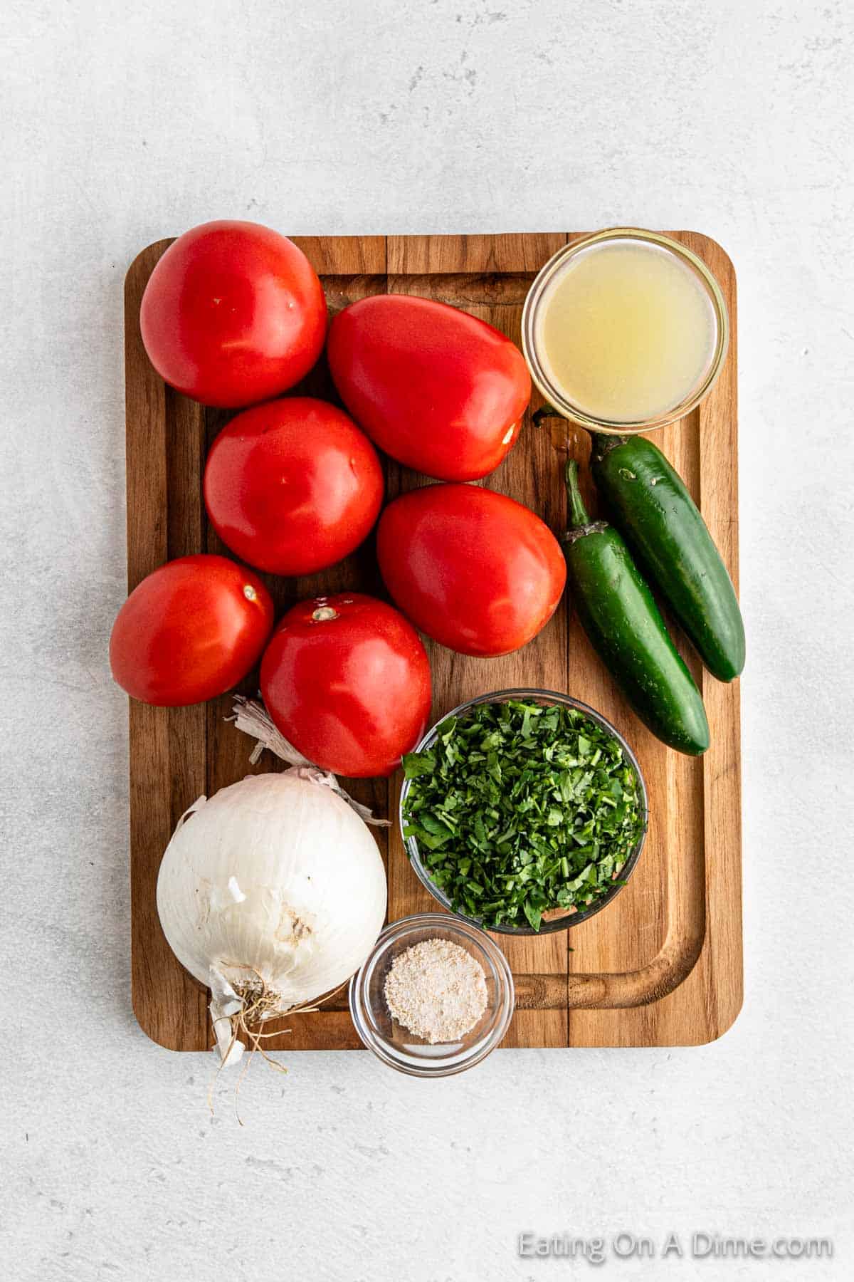 A wooden cutting board showcases the essentials for easy Pico De Gallo: a bunch of tomatoes, two jalapeños, a whole white onion, chopped cilantro in a bowl, salt in another bowl, and a glass bowl filled with lemon juice.