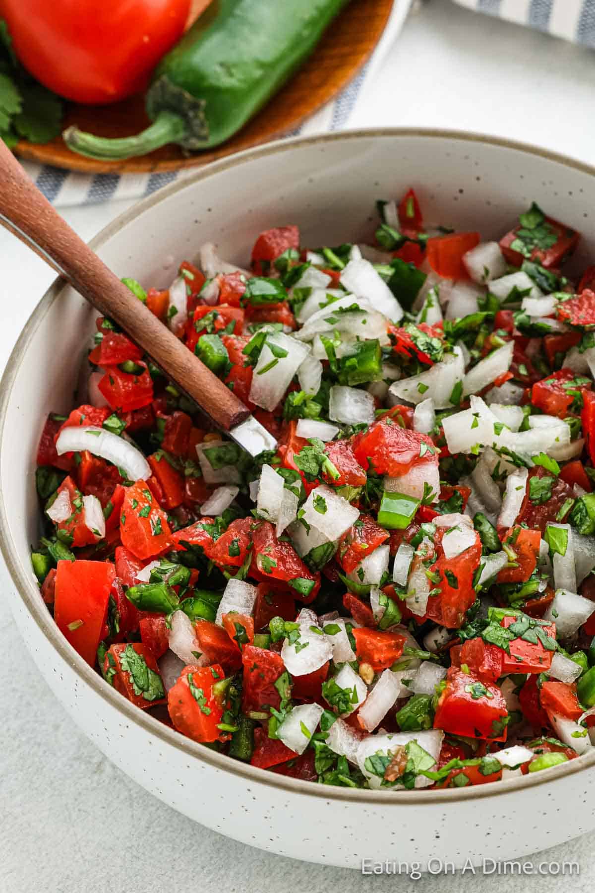 A bowl of fresh, easy Pico De Gallo made with chopped tomatoes, onions, jalapeños, and cilantro. A wooden spoon rests in the bowl. In the background, a plate holds a whole tomato and a jalapeño.