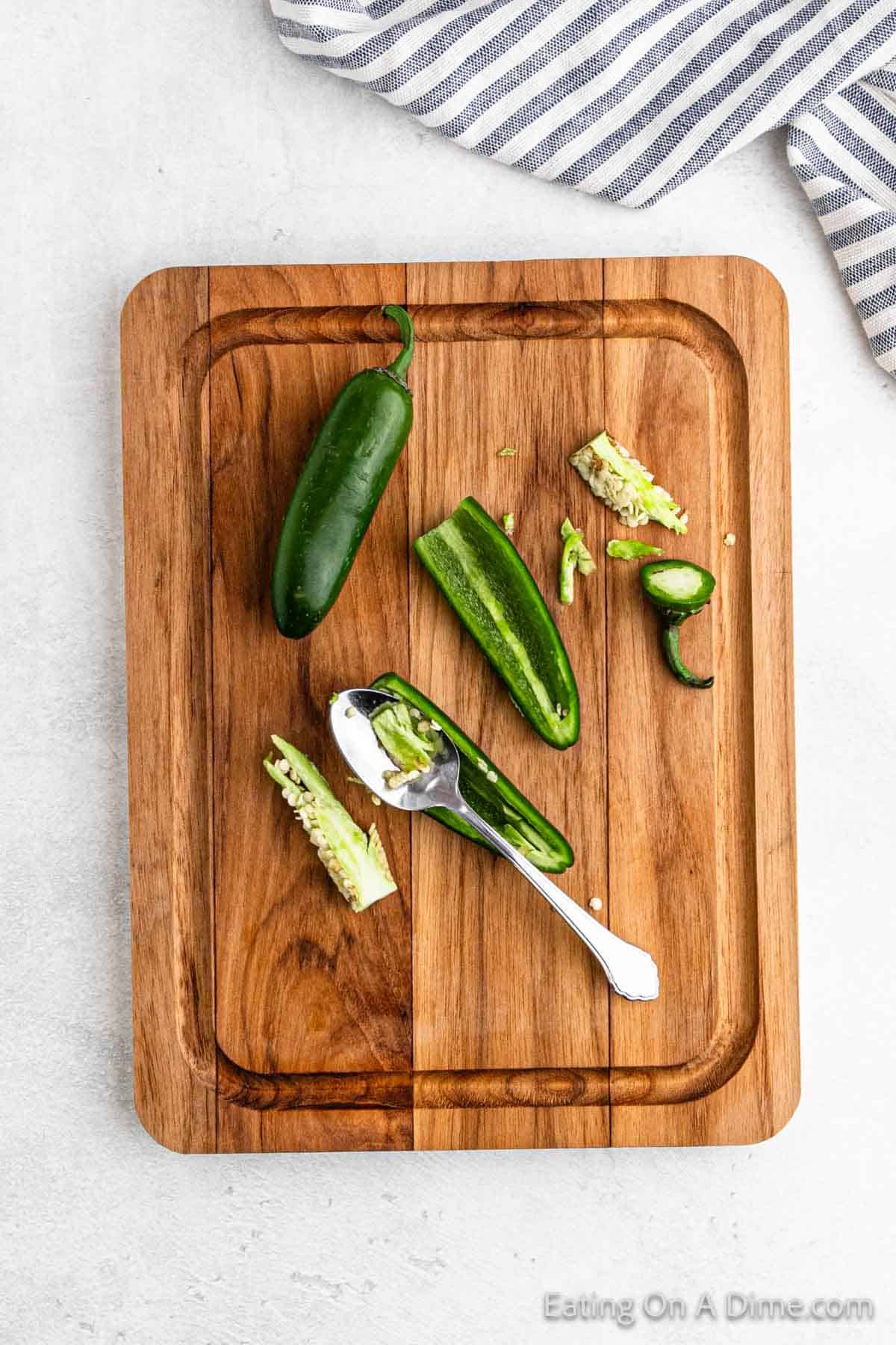 Two whole jalapeños and a halved one rest on a wooden cutting board, ready for an easy Pico De Gallo. One half is being seeded with a spoon, scattering seeds around. A striped cloth is partially visible at the top right corner.