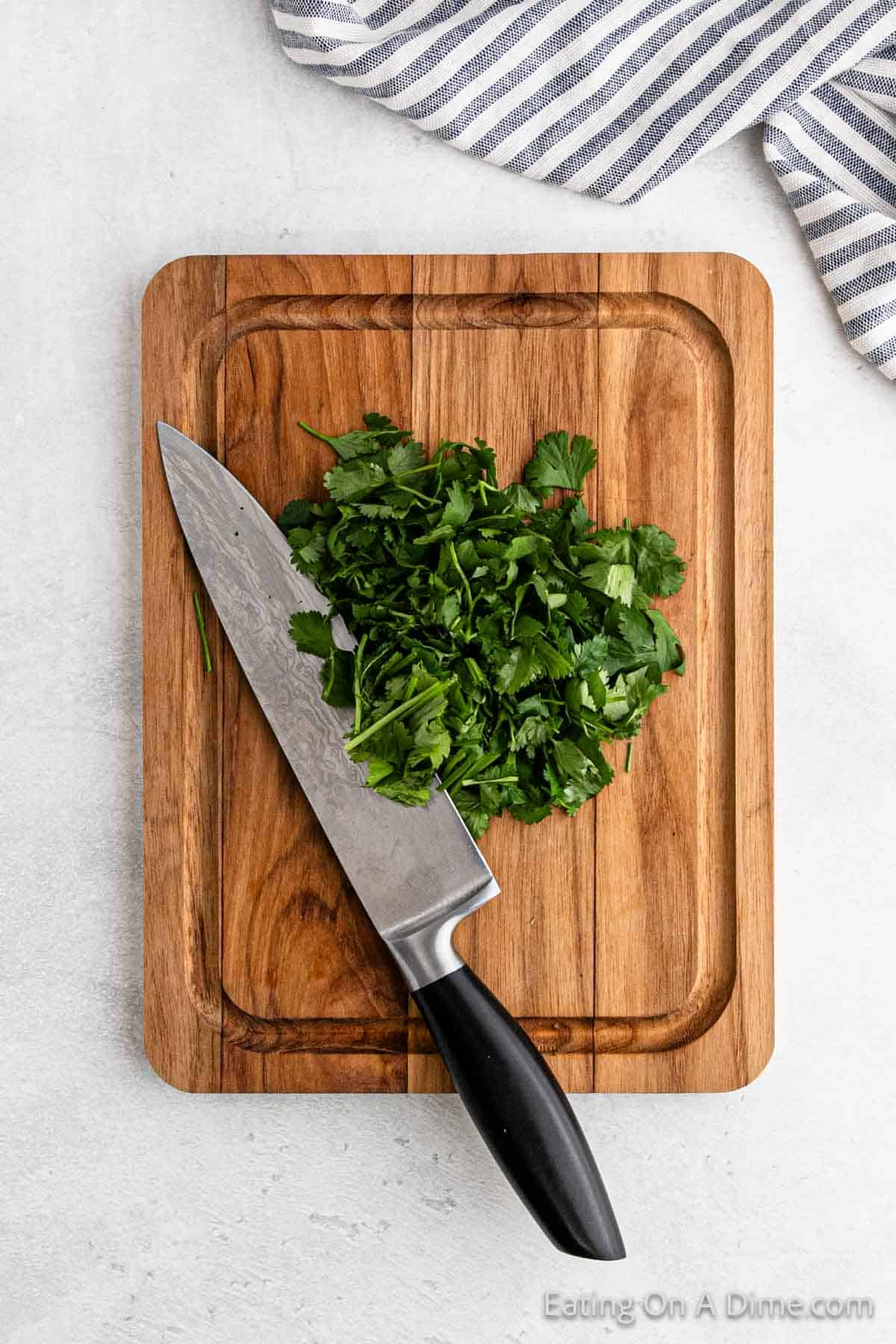 Chopped cilantro on a wooden cutting board with a chef's knife next to it, ready for an easy Pico De Gallo. A blue and white striped cloth is partially visible in the corner.