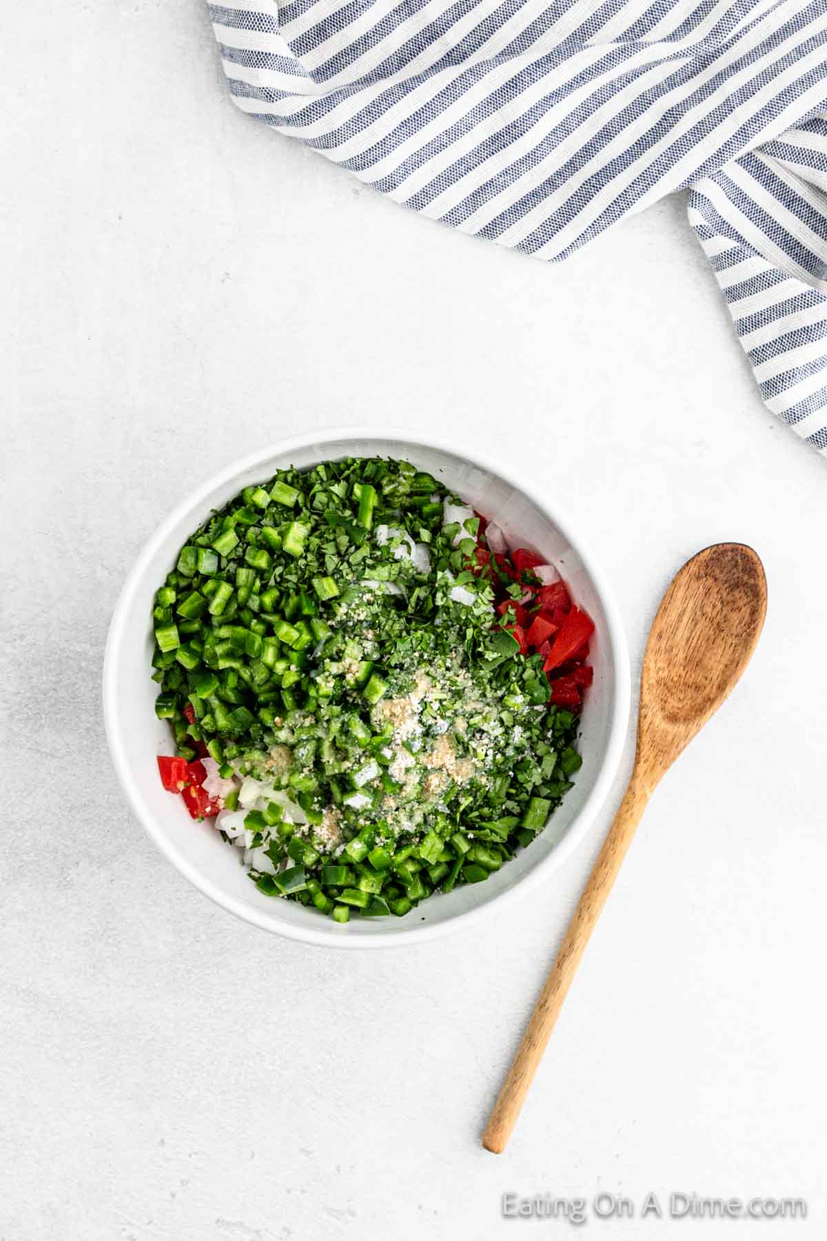 A white bowl filled with chopped green and red vegetables, reminiscent of easy Pico De Gallo, is paired with fresh herbs and grated cheese. A wooden spoon rests beside the bowl on a light surface, while a striped cloth peeks from the corner.