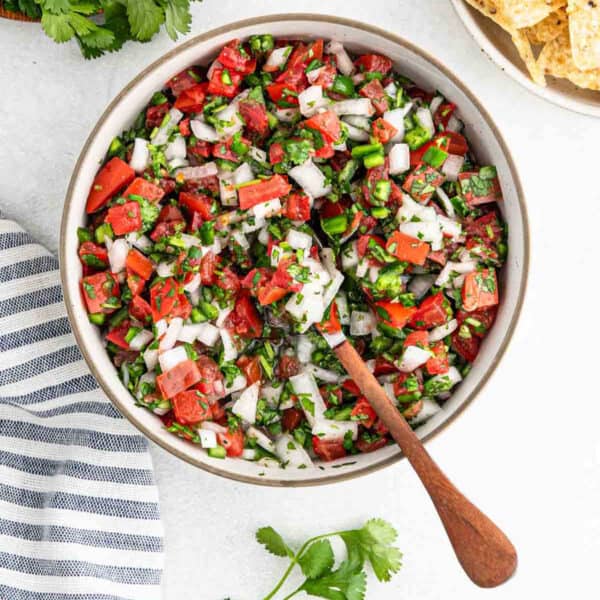 A bowl of vibrant easy pico de gallo sits on a table, featuring diced tomatoes, onions, cilantro, and jalapeños. A wooden spoon rests inside, while a striped napkin and tortilla chips are nearby. A sprig of cilantro adds the finishing touch to this delightful scene.