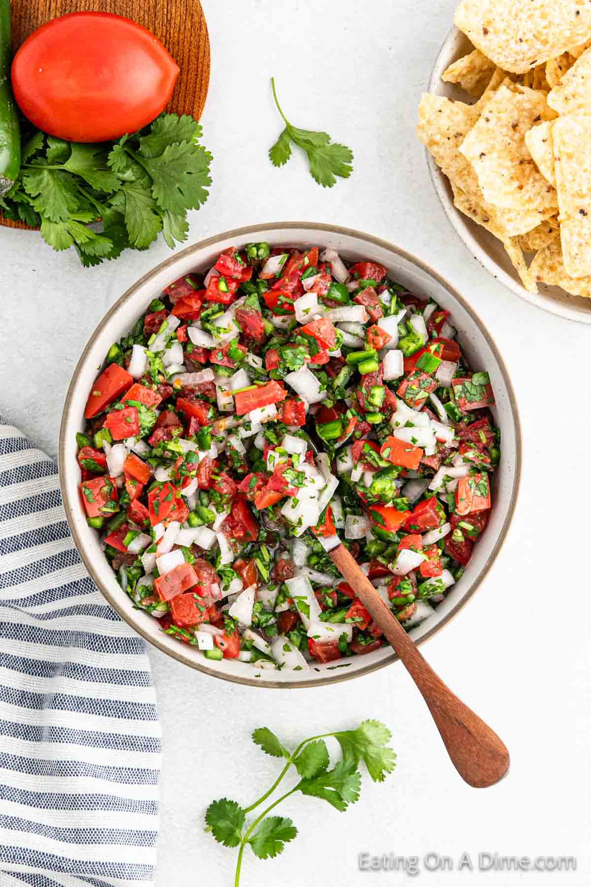 A bowl of easy Pico De Gallo with diced tomatoes, onions, cilantro, and jalapeños sits on a table. Nearby, tortilla chips await alongside a whole tomato, fresh cilantro leaves, and a blue-striped napkin.