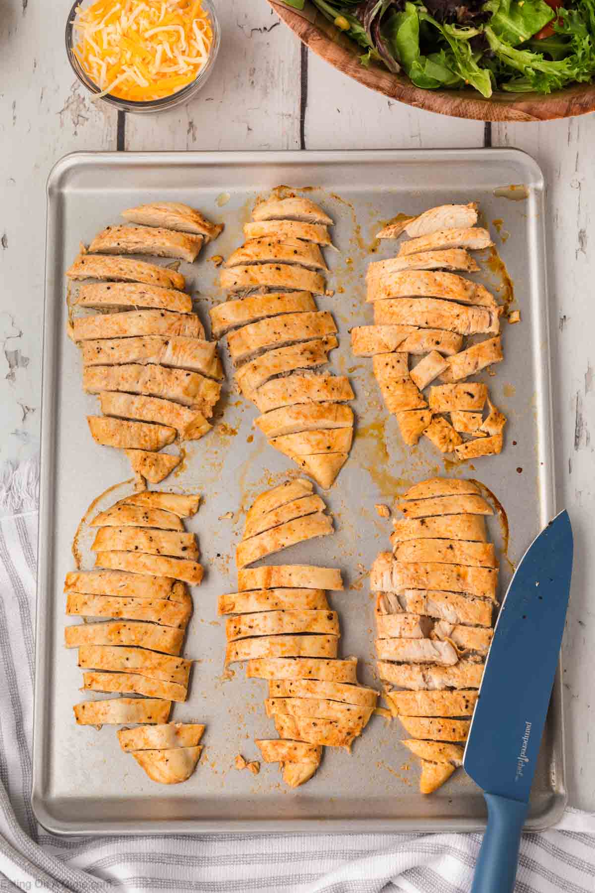 Sliced cooked chicken breasts rest on a baking sheet beside a blue knife, ready to transform into a delicious Chick-Fil-A Southwest Salad. Bowls of shredded cheese and mixed greens await in the background, promising layers of flavor and texture.