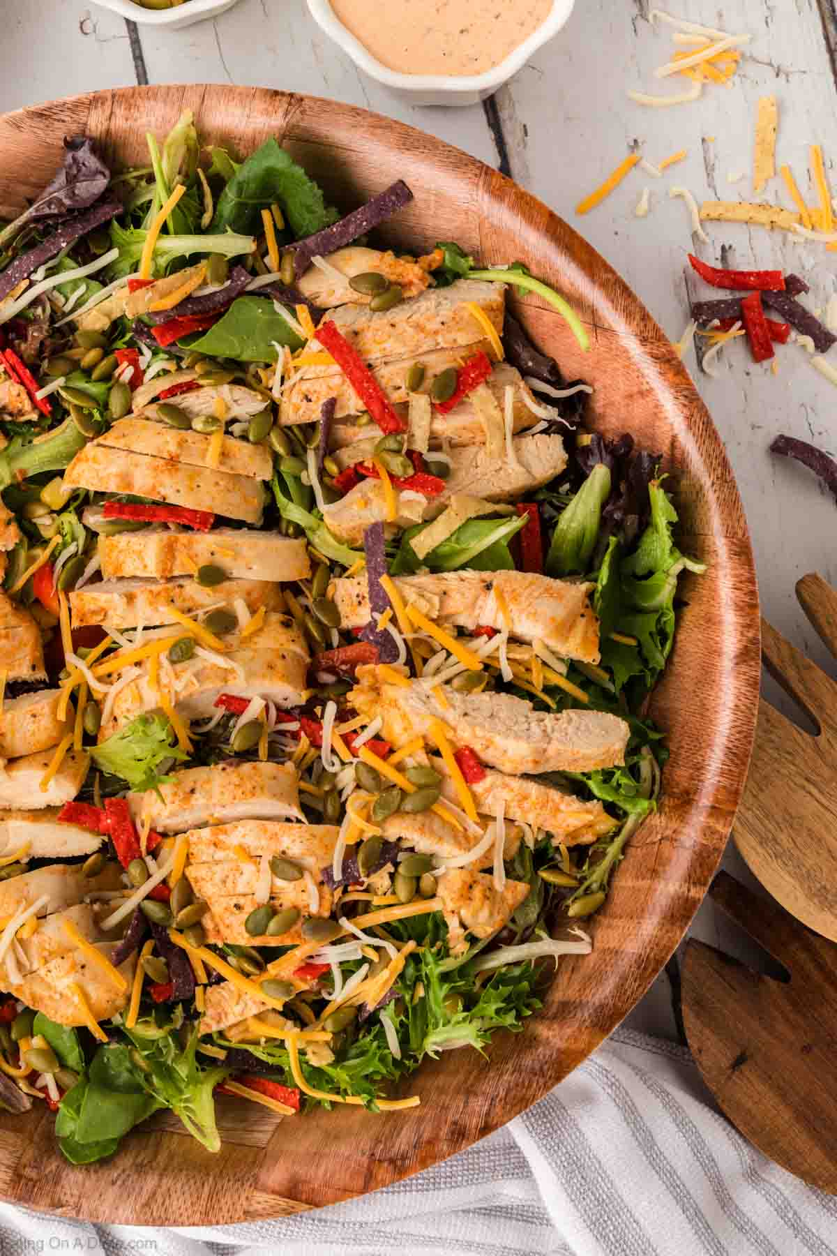 A wooden bowl brimming with a Chick-Fil-A Southwest Salad, showcasing grilled chicken slices atop mixed greens, shredded cheese, red tortilla strips, and pumpkin seeds. A wooden serving spoon is nearby and a small container holds the signature creamy dressing.