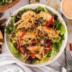 A vibrant Chick-Fil-A Southwest Salad featuring grilled chicken slices, shredded cheese, sliced red peppers, corn, seeds, and a mix of leafy greens, served in a bowl. The background includes a striped napkin and cutlery.