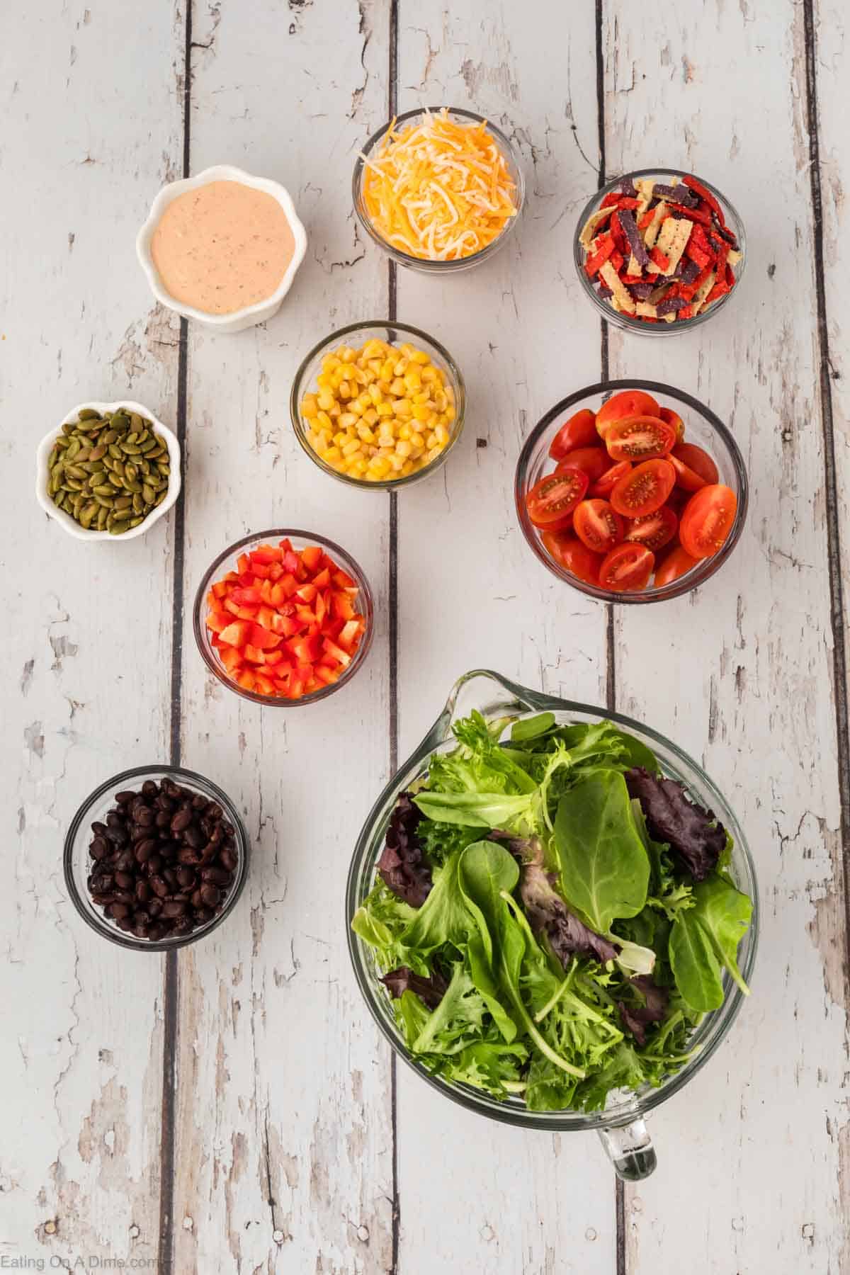 A rustic display of Chick-Fil-A Southwest Salad ingredients: mixed greens in a large bowl, surrounded by small bowls of cherry tomatoes, corn, shredded cheese, tortilla strips, diced red peppers, black beans, pepitas, and a creamy dressing.