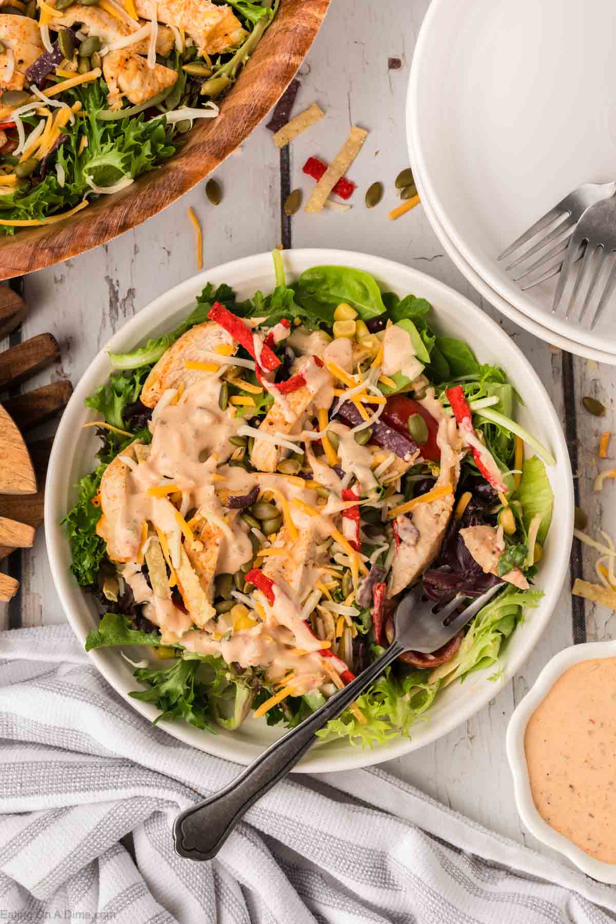 A bowl of Chick-Fil-A Southwest Salad featuring mixed greens, grilled chicken, shredded cheese, sliced red bell peppers, corn, and tortilla strips, drizzled with a creamy dressing. A fork rests in the bowl beside a small dish of dressing on a wooden table.