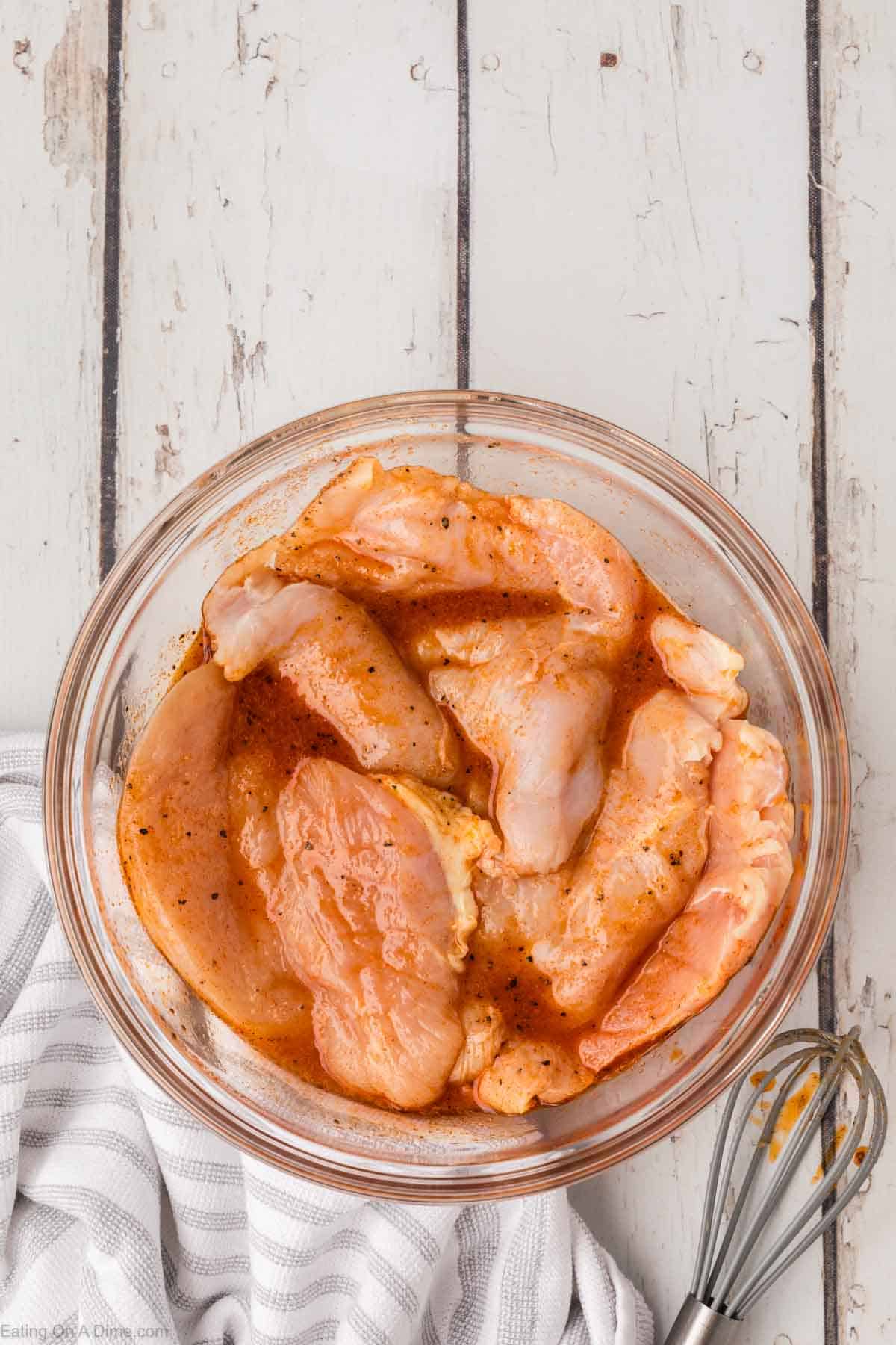 A glass bowl filled with raw, seasoned chicken breast fillets sits ready for a dish as delightful as a Chick-Fil-A Southwest Salad. On the whitewashed wooden surface, a striped kitchen towel and a small whisk add to the preparation scene.