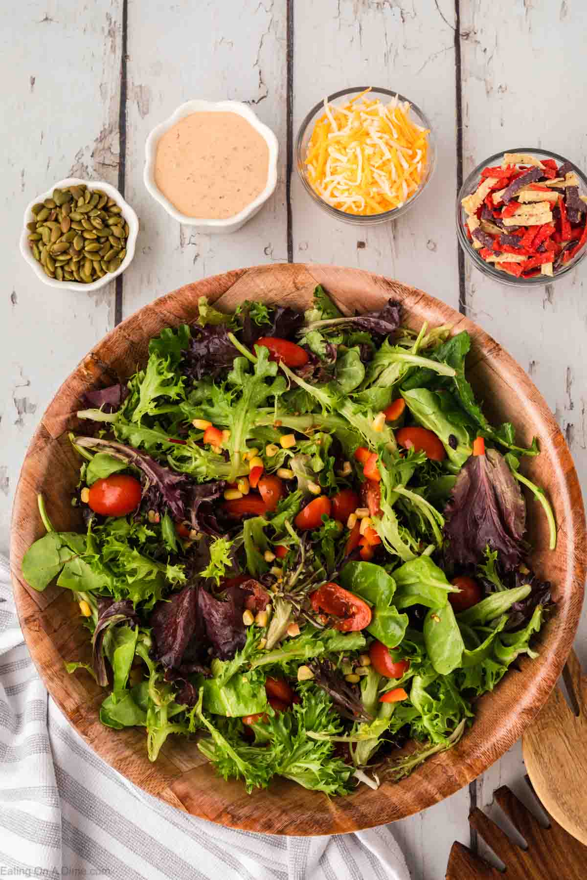 A wooden bowl brimming with a Chick-Fil-A Southwest Salad, featuring mixed greens, cherry tomatoes, and seeds. Surrounding the bowl are small dishes of creamy dressing, shredded cheese, tortilla strips, and pumpkin seeds on a rustic wooden table.