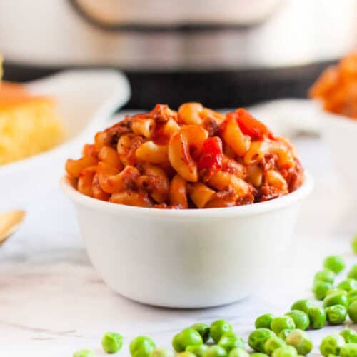 A white bowl brimming with Instant Pot macaroni and beef chili, garnished with vibrant tomato bits. Sitting on a marble surface, green peas are scattered in the foreground. In the background, hints of other dishes nod to an American goulash recipe tradition.