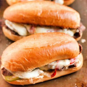 Close-up of two toasted Philly cheesesteak sandwiches, brimming with melted cheese, thinly sliced beef, and sautéed red and green peppers. The mouthwatering creations rest enticingly on a baking tray, ready to satisfy any craving.