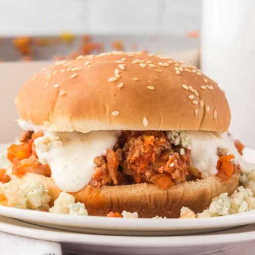 A Buffalo chicken sloppy Joe sandwich topped with creamy sauce and crumbled blue cheese on a sesame seed bun, served on a white plate. The filling looks hearty with visible chunks of meat and vegetables.