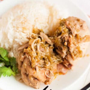 A plate featuring two pieces of crock pot salsa verde chicken topped with green chili sauce, accompanied by a serving of white rice. A sprig of cilantro decorates the side of the plate.