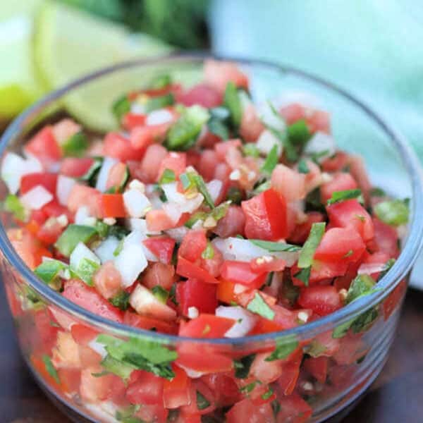 A glass bowl brimming with an easy pico de gallo recipe showcases a vibrant mix of chopped tomatoes, onions, jalapeños, and cilantro. Lime wedges are subtly visible in the background.