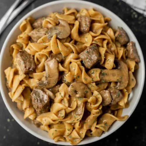 A bowl of creamy beef stroganoff with wide egg noodles, chunks of beef, and sliced mushrooms, garnished with chopped herbs, makes for an easy crock pot recipe.