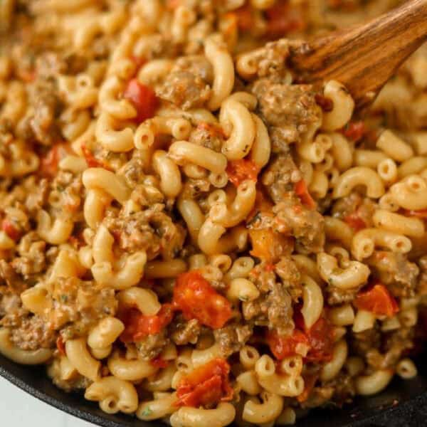 A close-up of a skillet containing macaroni pasta mixed with ground beef, diced tomatoes, and savory sausage. A wooden spoon is stirring the ingredients, showcasing this hearty twist on a classic macaroni and cheese recipe.