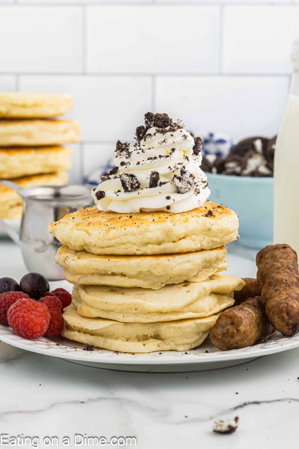 Oreo Pancakes stacked on a plate with a side of sausage links, fresh raspberries and grapes topped with whipped cream and crushed Oreos