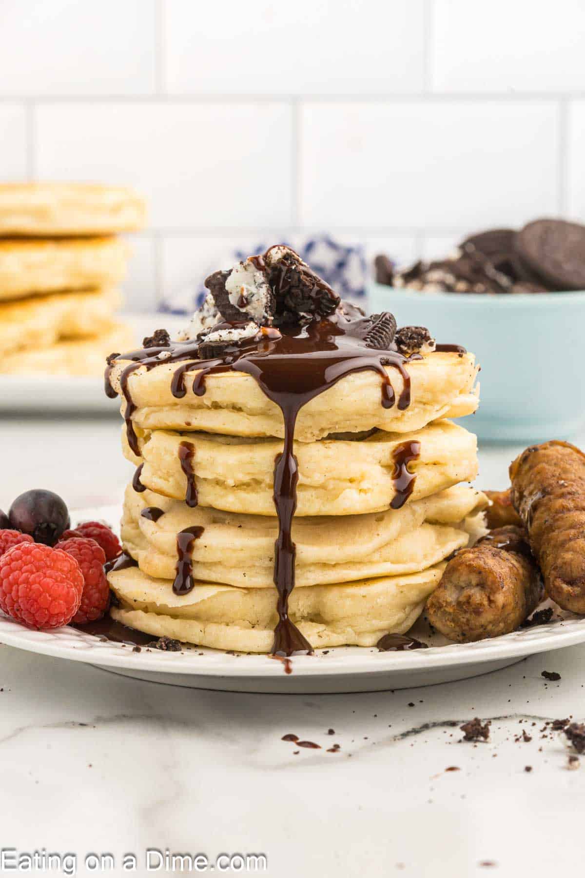Oreo pancakes stacked on a plate topped with chocolate sauce and crushed oreos with a side of fresh raspberries and grapes