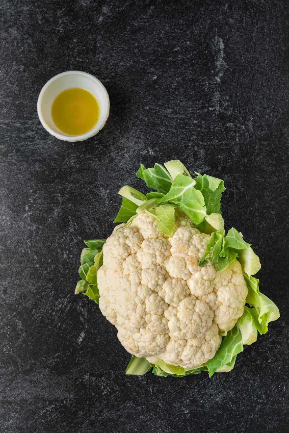 A head of cauliflower with green leaves rests on a dark, textured surface. Nearby, a small white bowl containing olive oil awaits the transformation process—perfect for those learning how to season cauliflower rice to perfection.