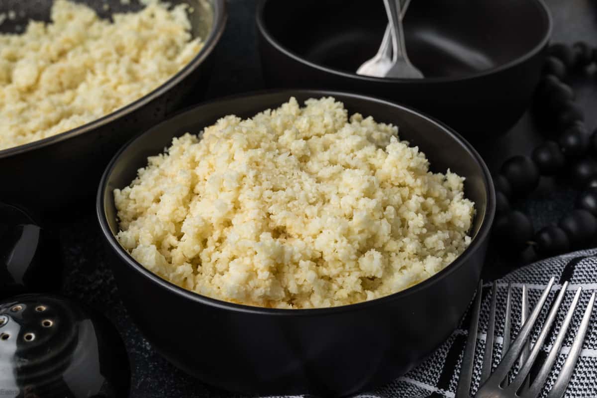 A black bowl filled with fluffy, cooked couscous sits on a dark surface. Two spoons rest in a smaller black bowl nearby, hinting at the next step: how to season cauliflower rice. A black and white patterned cloth and black beaded decor adorn the background elegantly.