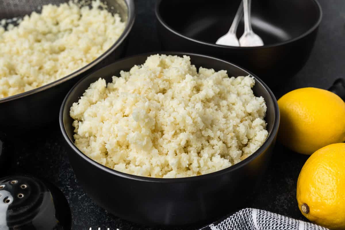 On a dark countertop, a bowl holds fluffy, cooked cauliflower rice—perfect for discovering how to season cauliflower rice. Nearby, two whole lemons rest beside a black-and-white checkered cloth. In the background, two black bowls sit ready with spoons.