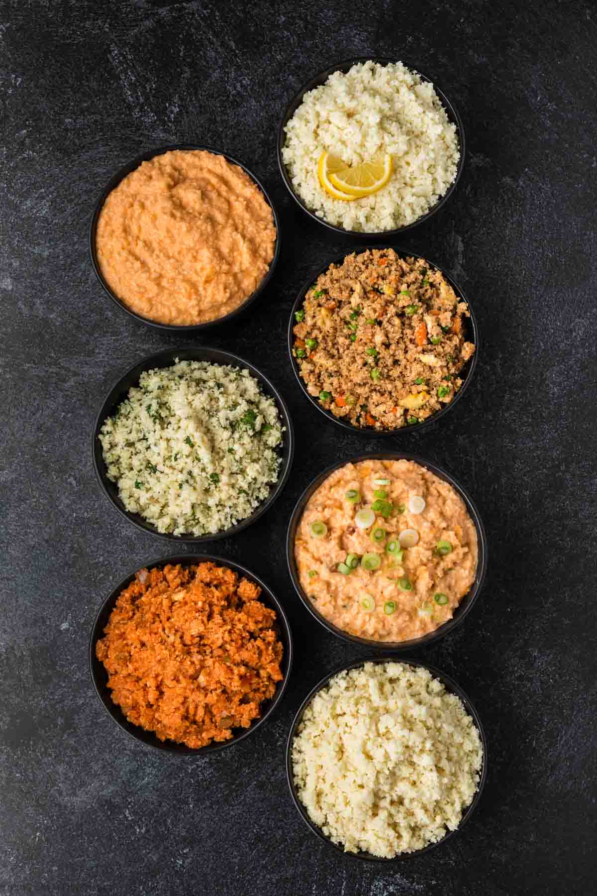 Seven bowls of cauliflower rice, seasoned to perfection, with various toppings and flavors like tomato, herbs, and mixed vegetables, are arranged on a dark surface. For those wondering how to season cauliflower rice, one bowl showcases lemon slices as a fresh garnish.