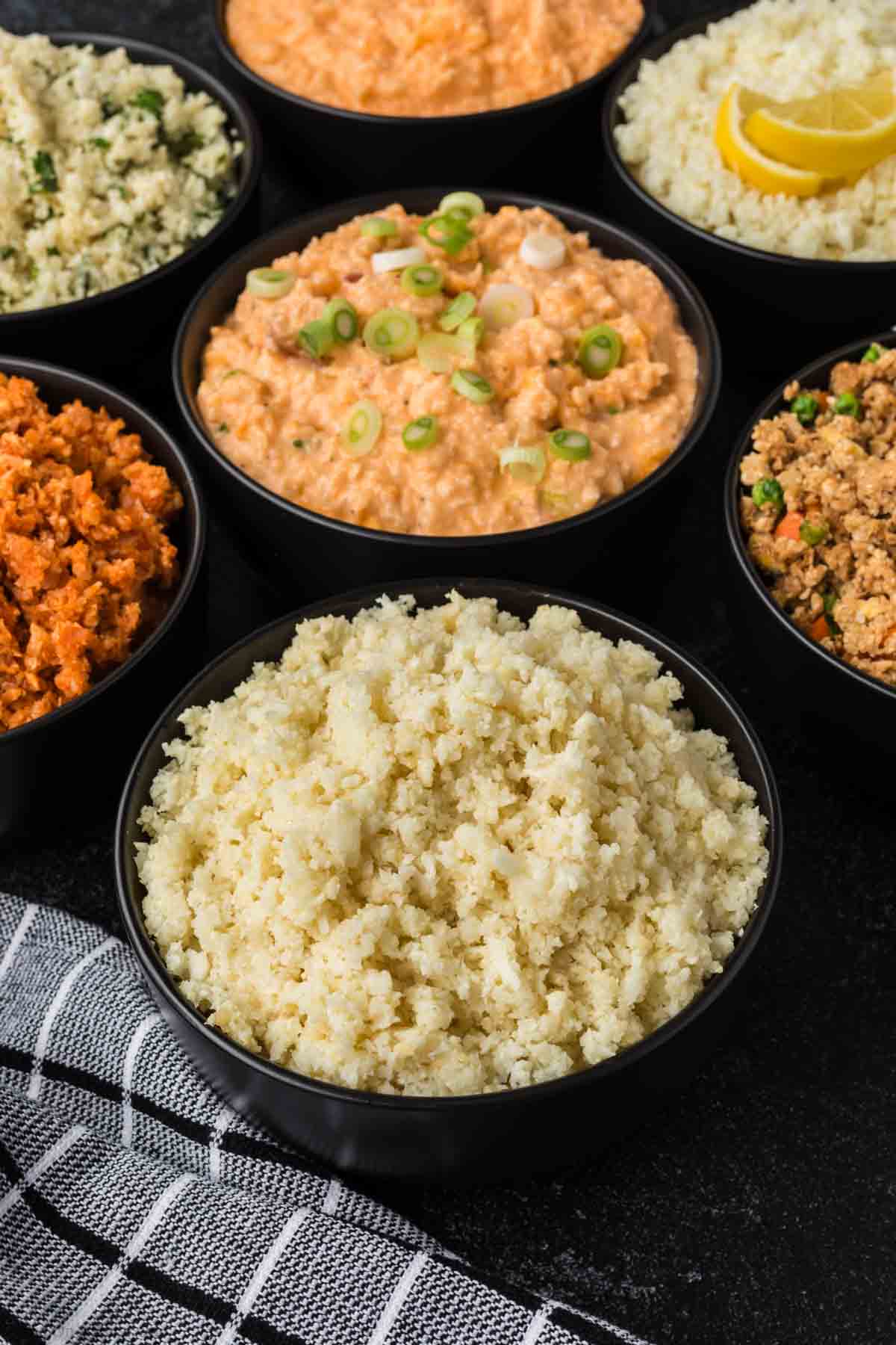 A variety of small bowls filled with different flavored cauliflower rice—perfect for discovering how to season cauliflower rice—are arranged in a circle. The flavors include plain, orange-colored, and garnished with green onions. A checkered cloth is partially visible at the bottom.