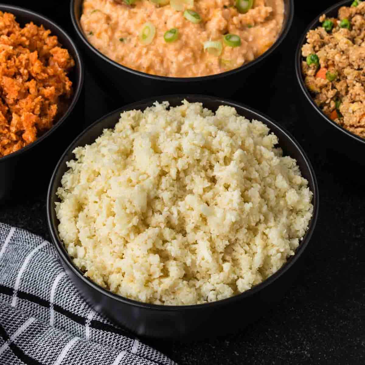 A bowl of perfectly seasoned cauliflower rice is surrounded by three other bowls containing different dishes. A black and white checkered cloth is partially visible next to the bowls on a dark surface, adding a touch of elegance to this guide on how to season cauliflower rice.