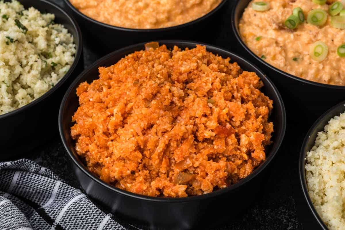 A close-up of bowls filled with different varieties of seasoned rice and grains. A vibrant orange-colored dish fills the central bowl, while others feature green herbs and garnishes, offering a glimpse of how to season cauliflower rice. A checkered cloth is partly visible in the foreground.