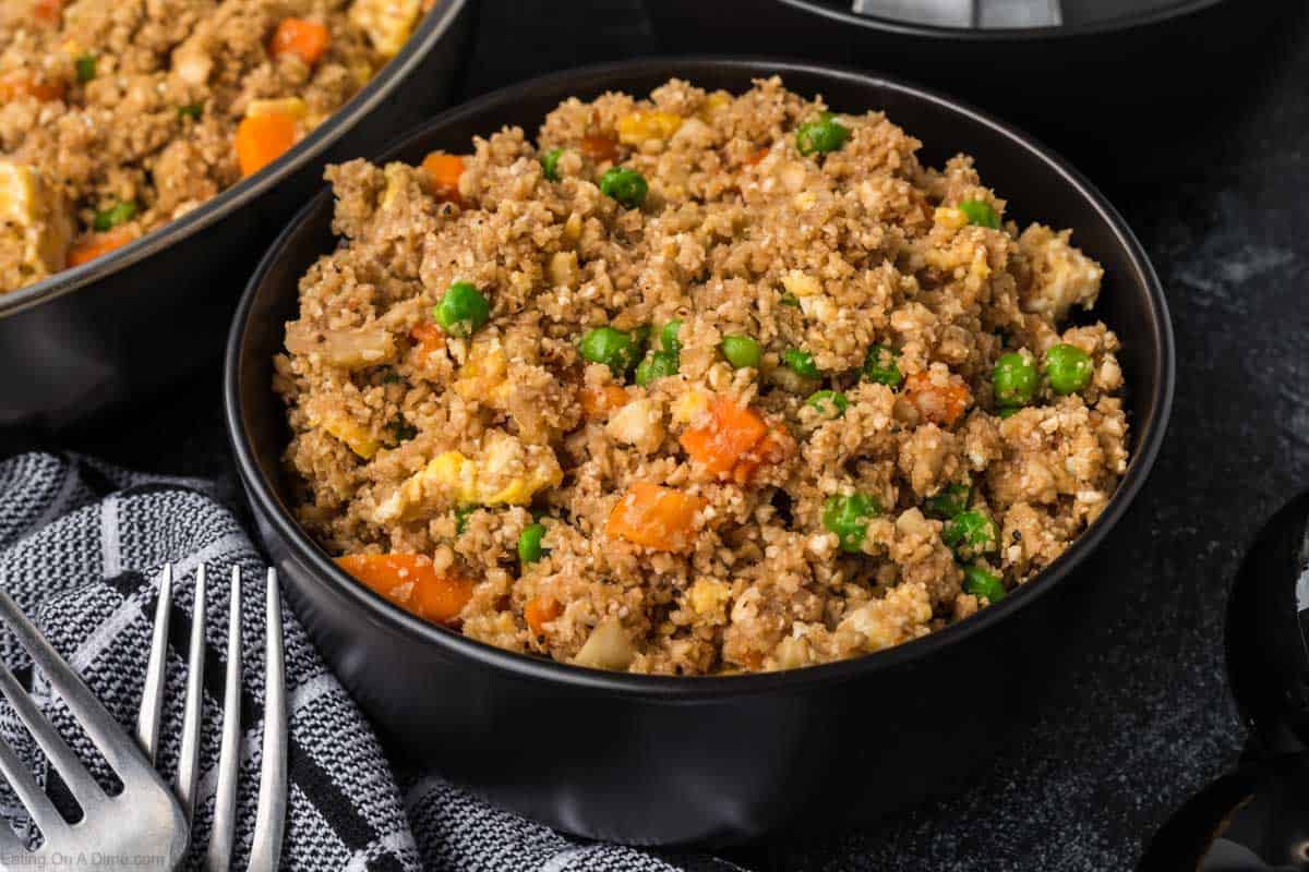 A black bowl brimming with fried rice features a delightful mix of peas, carrots, scrambled eggs, and perfectly seasoned cauliflower rice. The dish sits elegantly on a dark surface beside forks and a striped cloth napkin, offering a masterclass in how to season cauliflower rice expertly.