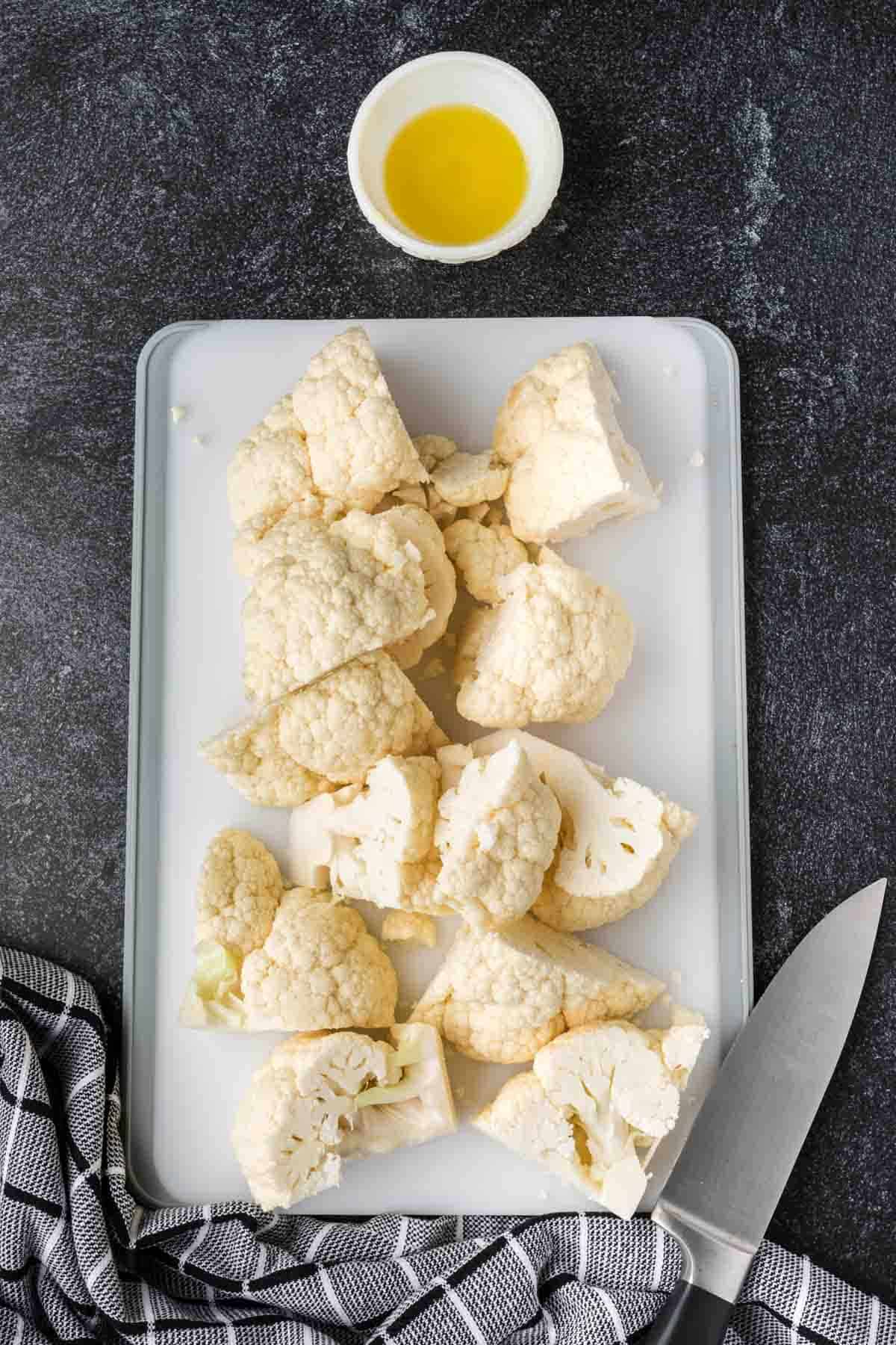 Chopped cauliflower florets on a white cutting board with a small bowl of olive oil above them, showing you how to season cauliflower rice. A knife and a black-and-white checkered cloth rest beside the board on a dark countertop.