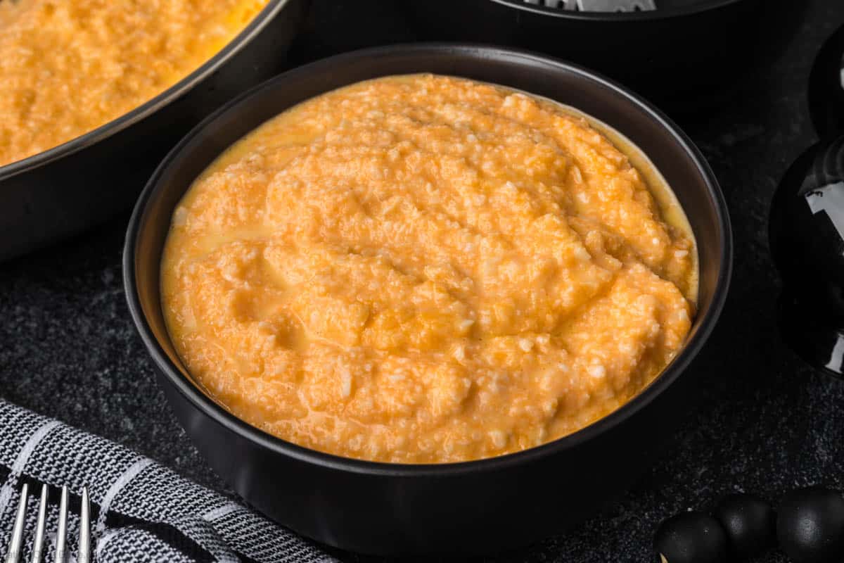 A black bowl brimming with creamy, golden-orange cheese grits rests on a dark surface. Part of a textured towel peeks into view, reminiscent of exploring how to season cauliflower rice for added flavor and warmth.